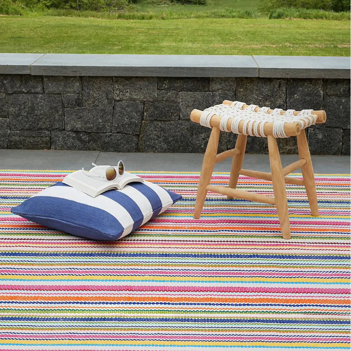 Dash and Albert Summer Stripe Handwoven Indoor Outdoor Rug laid outdoors with a stool, pillow, a book and sunglasses on top
