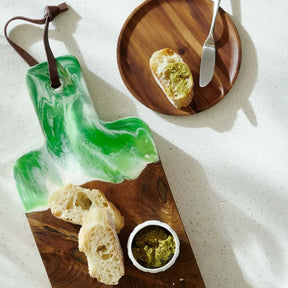 Blue Pheasant Austin Swirled Green Resin, Natural Teak Serving Board with two pieces of bread and dip on top in a room with a plat with food and spreader