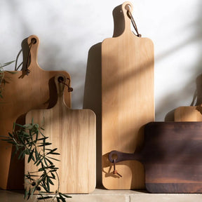 Blue Pheasant Edmund Natural Walnut Serving Board laid against the wall with other wood serving boards with a cast of shadows and greenry