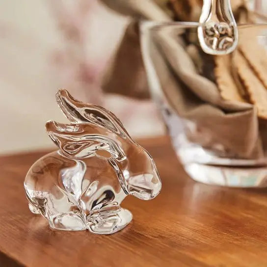 Simon Pearce Glass Rabbit set on a wood table in a room and next to a clear glass basket with a beige dinner napkin and crackers set inside the basket.