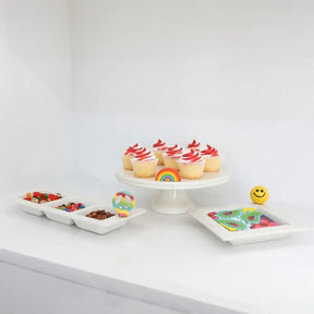 Nora Fleming Over the Rainbow Mini on a cake stand with cupcakes and set next to other trays with minis.