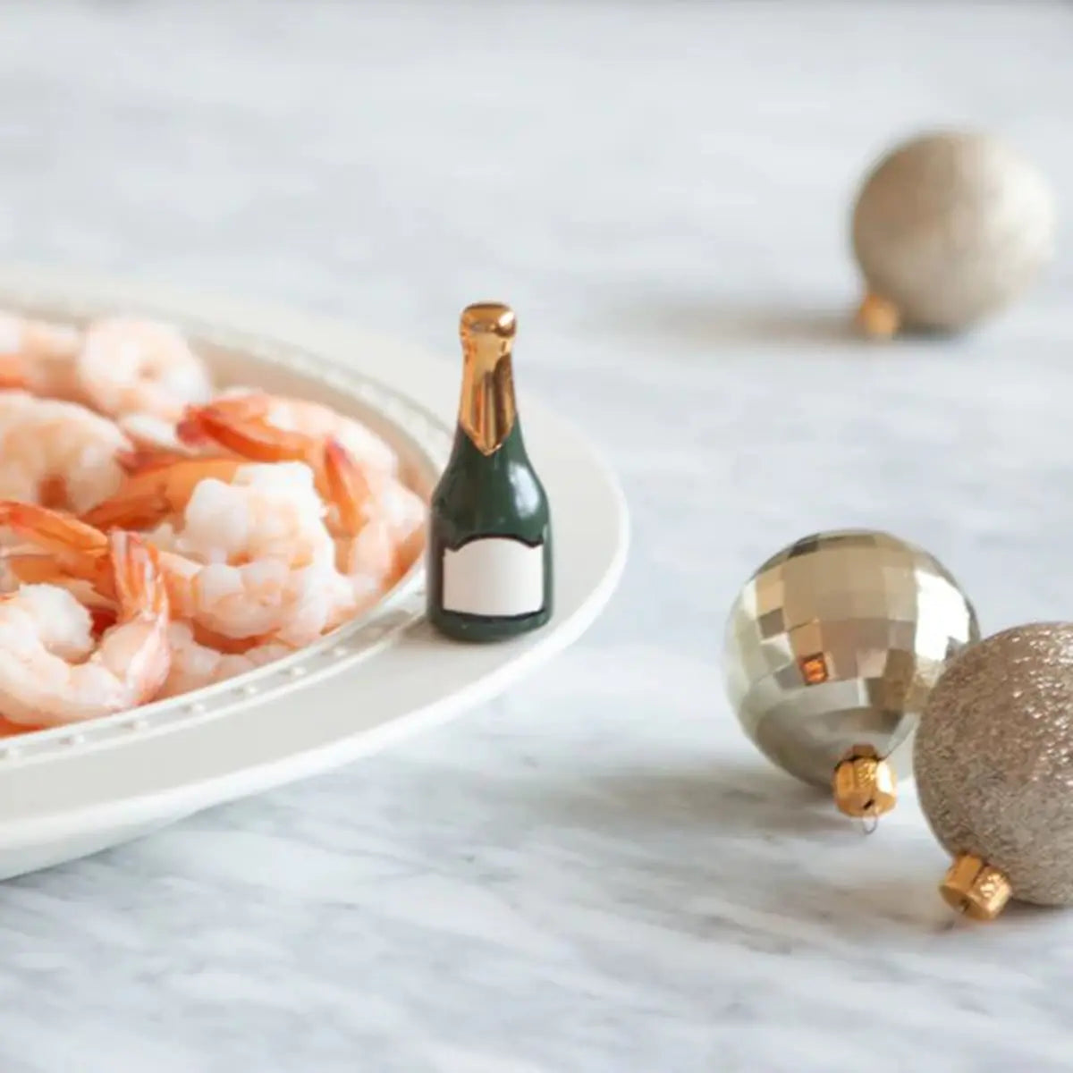 Nora Fleming Champagne Celebration Champagne Bottle Mini on a plate with cocktail shrimp with champagne colored ornaments laid on the table