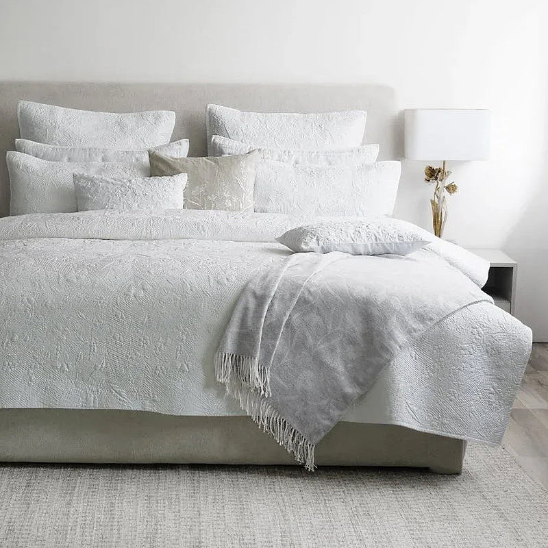 Image of bed with white textured Michael Aram bedding along with white rug and a white accent table with a white lamp