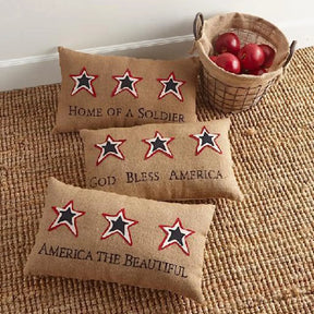 K and K Interiors Red Apple in a wire basket in a room with three decorative pillows with stars, reading HOME OF A SOLIER, GOD BLASS AMERICA, AMERICA THE BEAUTIFULE