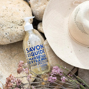 Compagnie de Provence Mediterranean Sea Liquid Soap laid on rocks and purple flowers and a sun hat