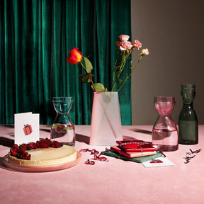 Mister and Missus Night Smoked Green Tall Water Set on a pink rug in a room with roses, a cheesecake with a velvet green curtain backdrop.