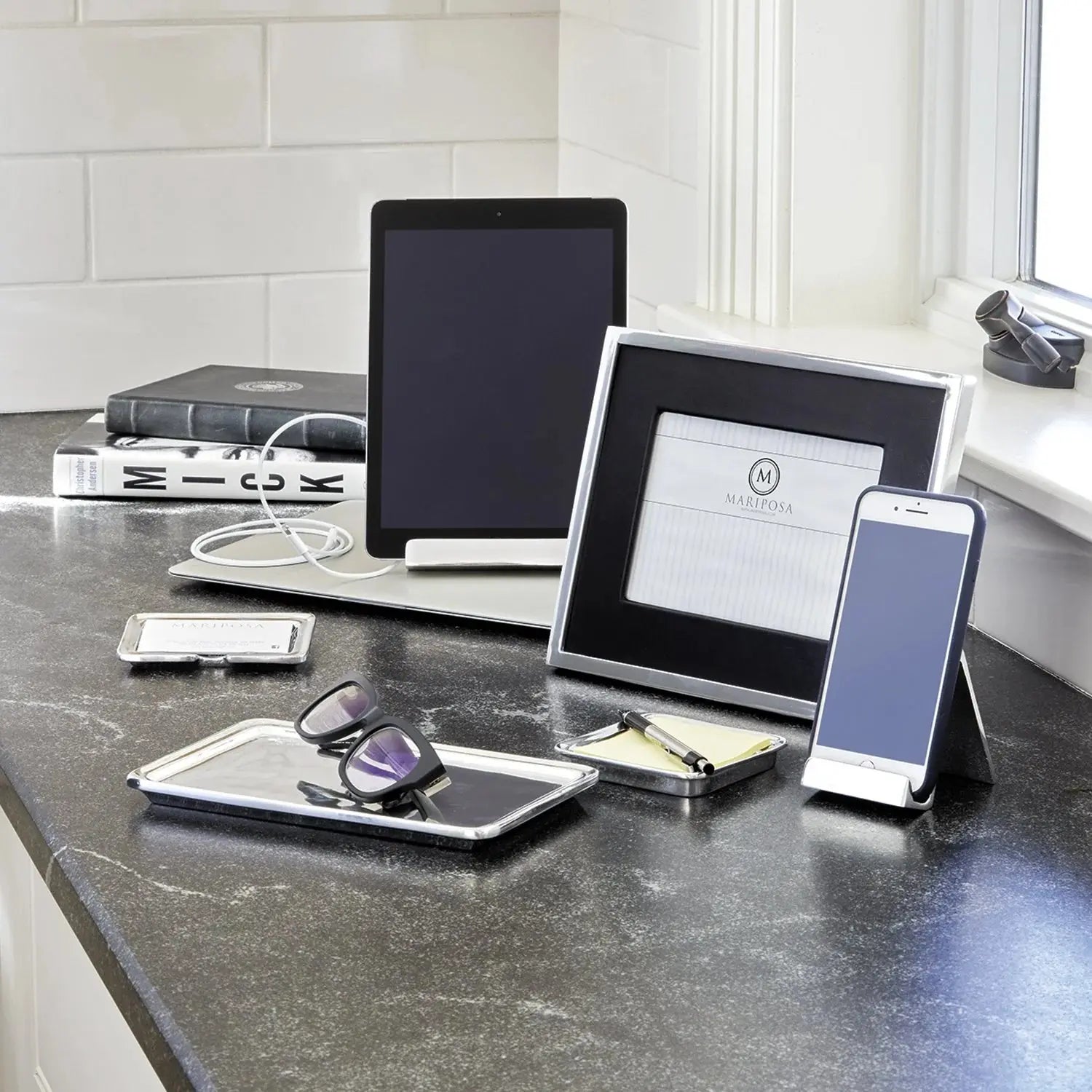 Mariposa Silver Signature Phone Holder with a phone set on a black marble desk, alongside a metal tray with glass, tablet and picture frame and books.