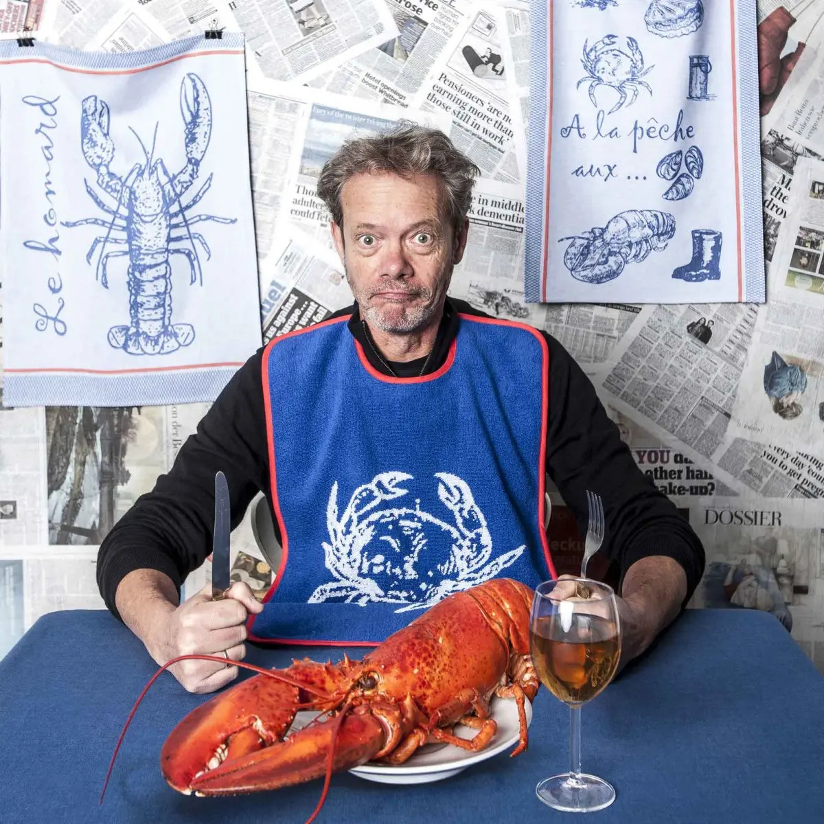 A man eatting with a lobster bib and Coucke A la Peche PJ Tea Towel hung on the wall