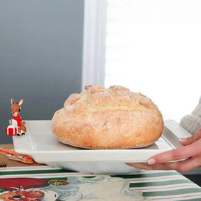 Nora Fleming Rudolph the Red-Nosed Reindeer Mini on a square plater in a room with a loaf of bread