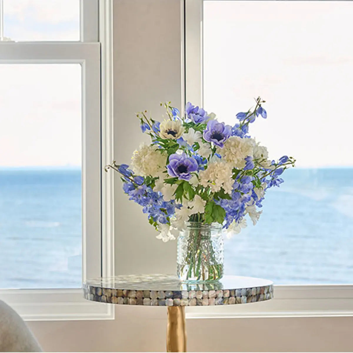 Diane James Blooms Blue Delphinium and Anemones in Vase set on a round table in a room with windows, showing the water and ocean.