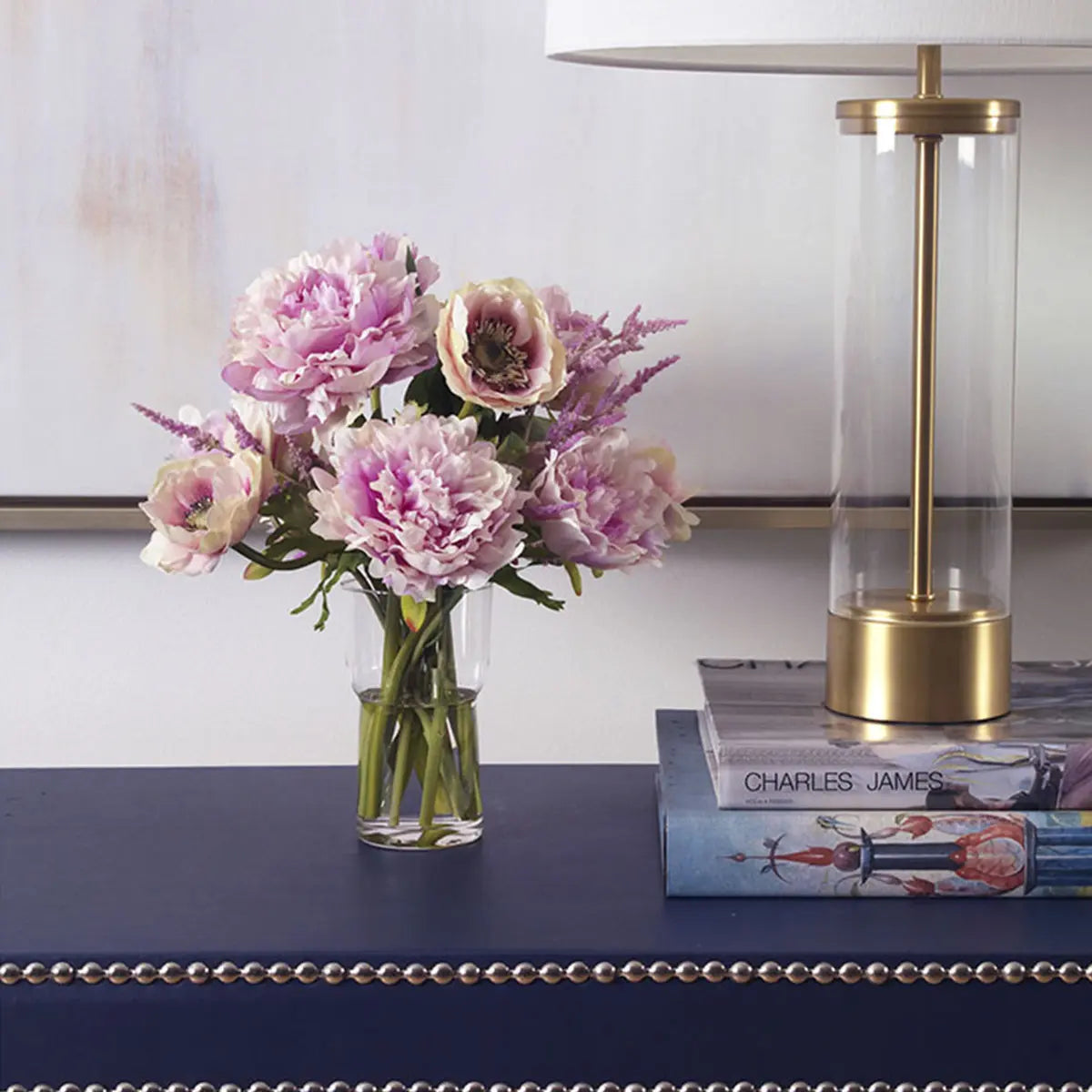 Diane James Pink Anemones and Peonies in Vase set on a navy blue console table by a lamp and books in a room