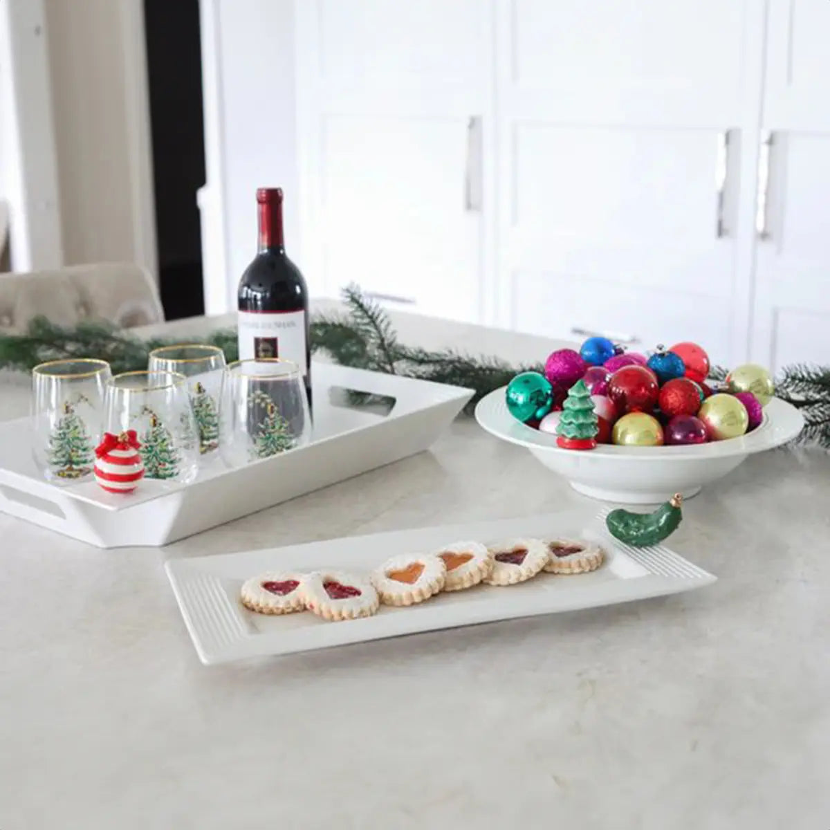 Nora Fleming Bread Tray with cookies on top with a pickle mini attached, set next to stemless wine glasses with christmas tree motifs and a wine bottle and small round ornaments in a bowl