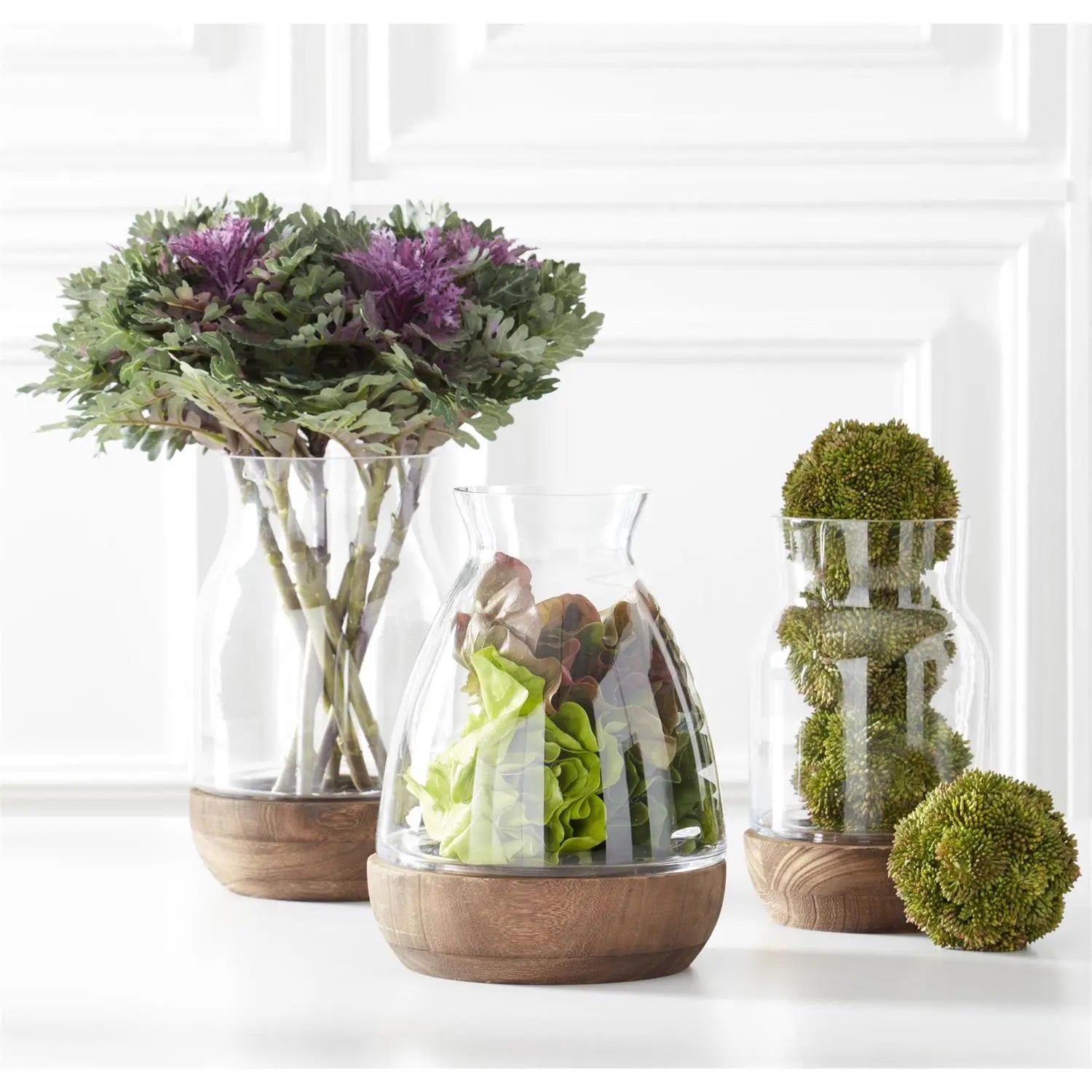 K and K Interiors Dark Green Sedum Ball stacked in a clear glass and one laid on the floor. Set next to other floral items